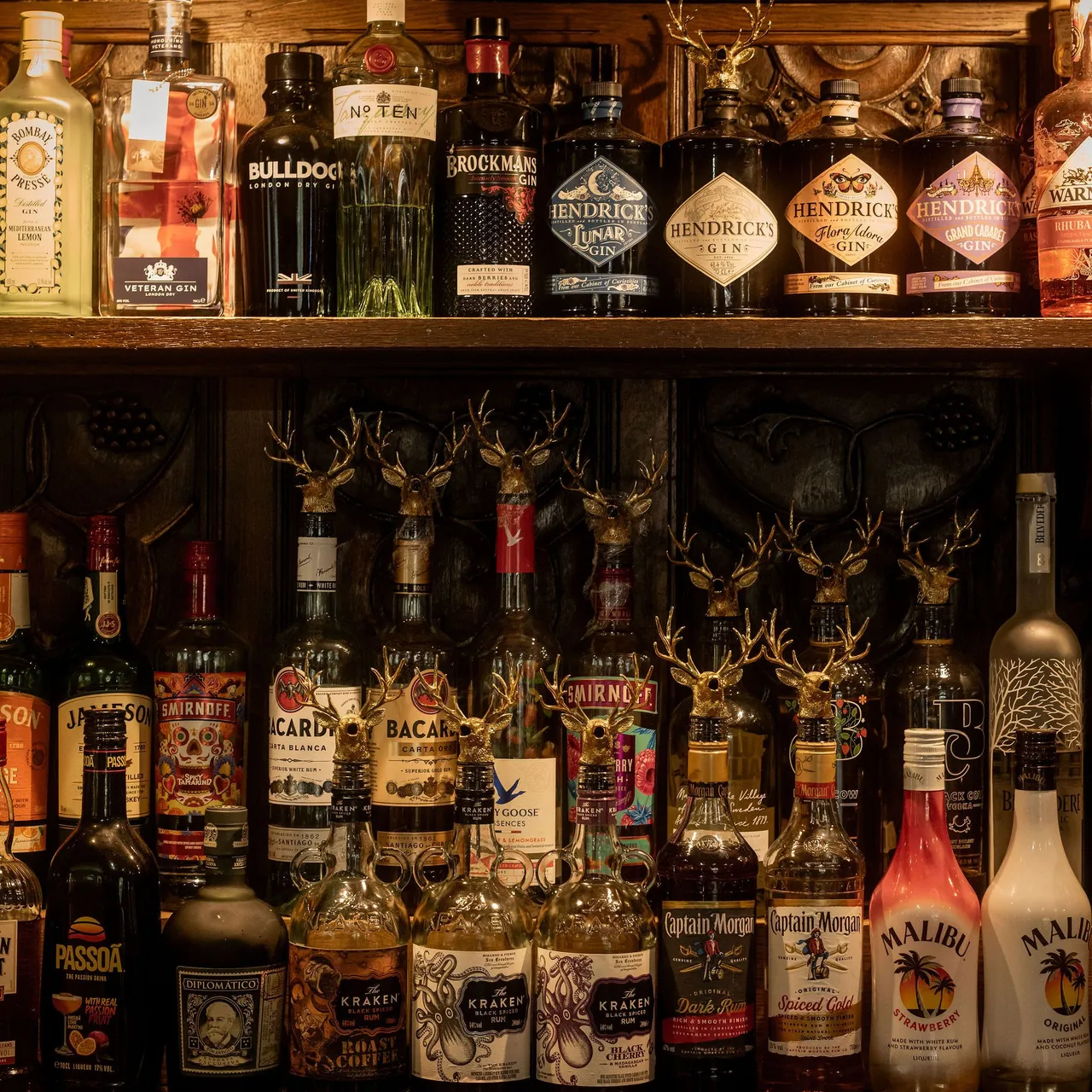 A well-stocked bar shelf featuring various bottles of liquor including gin, rum, vodka, and liqueurs, illuminated in a warmly lit setting.