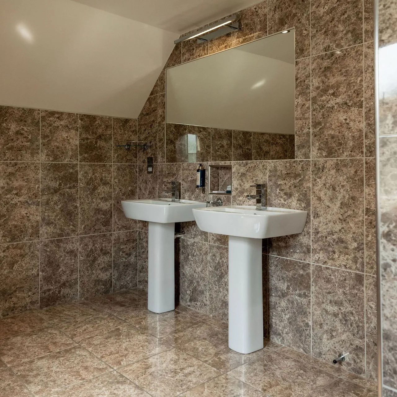 Modern bathroom with brown marble tiles, a large wall mirror, and two white pedestal sinks.