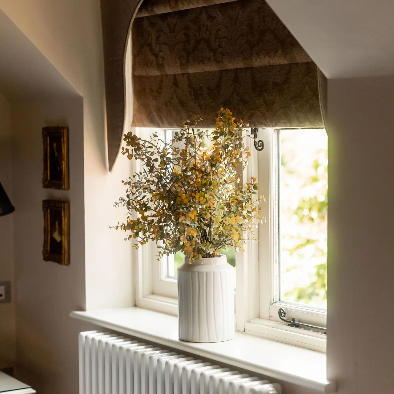 A vase with yellow flowers placed on a windowsill in front of a window with brown roman shades, above a white radiator.