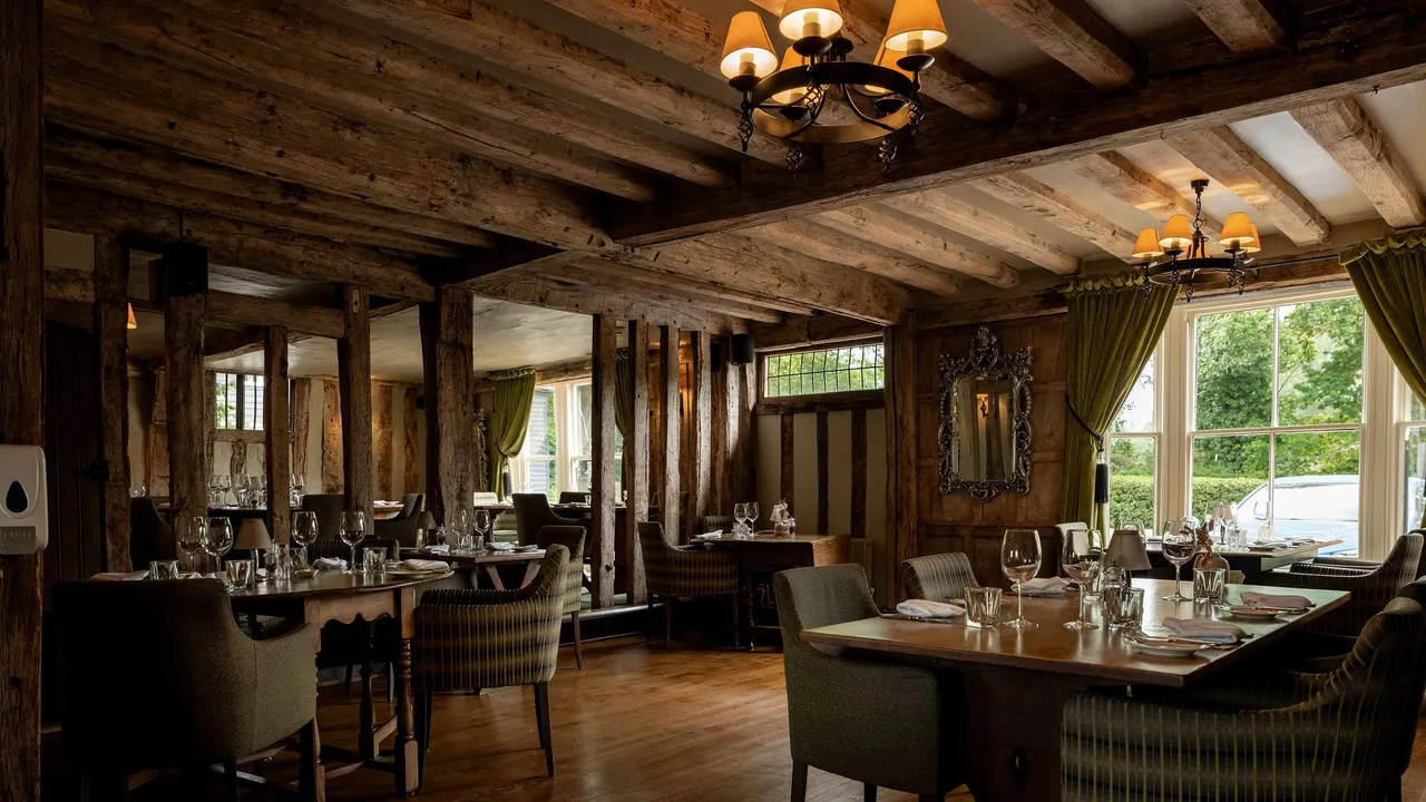 Cozy restaurant interior with rustic wooden beams, elegant lighting, green curtains, and tables set with glassware and napkins near large windows.
