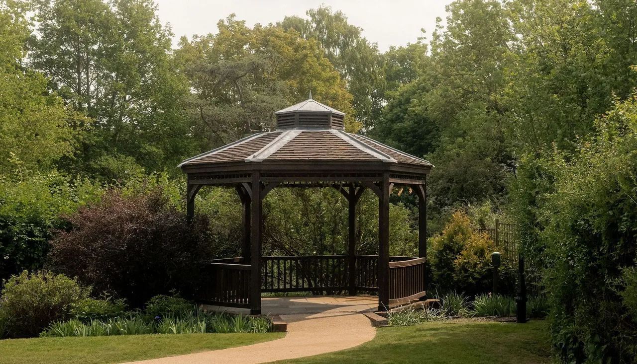 A wooden gazebo surrounded by greenery and trees, with a curved path leading to it in a garden setting.