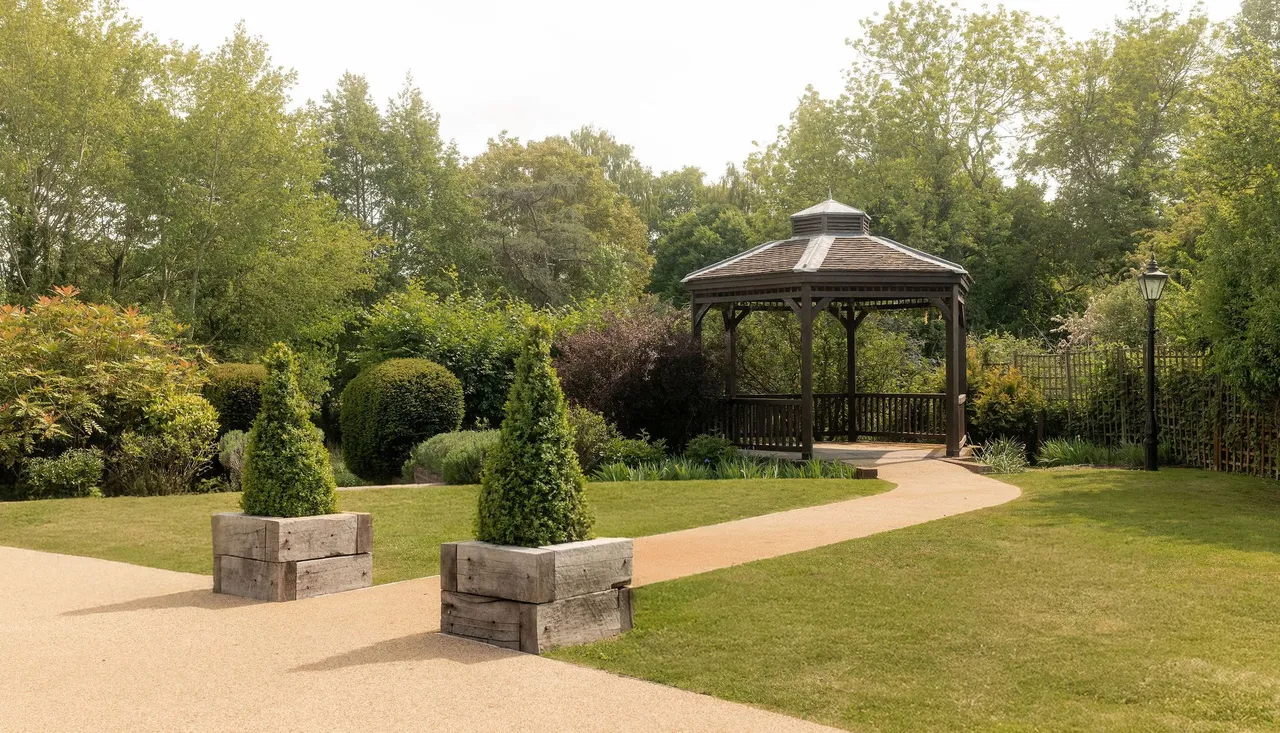 A peaceful garden with a wooden gazebo, neatly trimmed hedges, green trees, a paved pathway, and decorative planter boxes with conical shrubs.