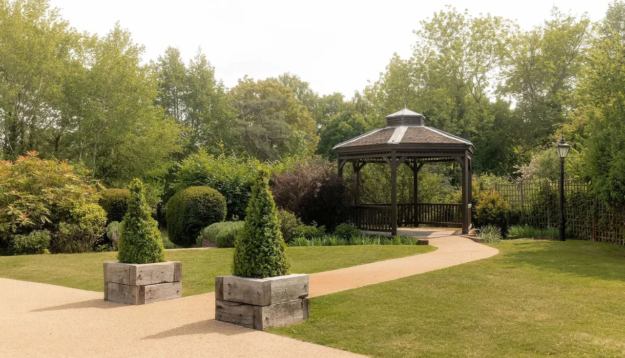 A peaceful garden with a wooden gazebo, neatly trimmed hedges, green trees, a paved pathway, and decorative planter boxes with conical shrubs.
