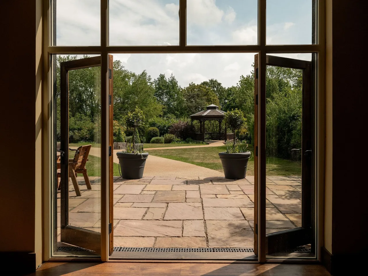 Open glass doors leading to a stone patio with two large potted plants and a view of a garden gazebo surrounded by greenery.