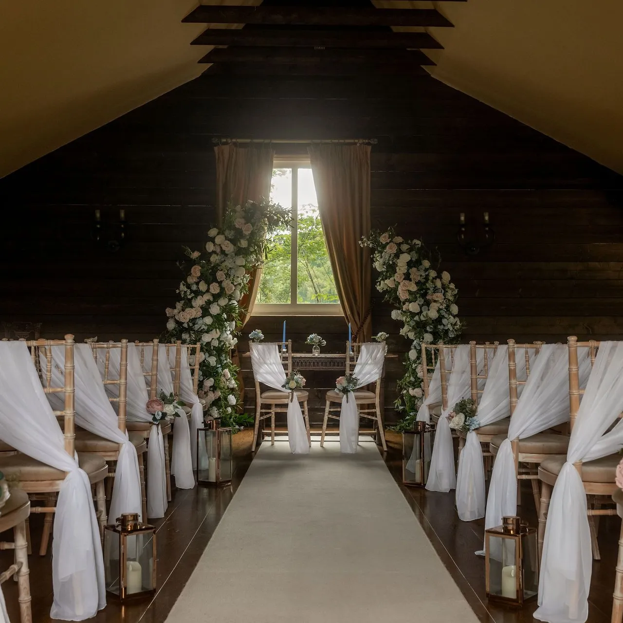 Rustic indoor wedding ceremony setup with floral arch and draped chairs