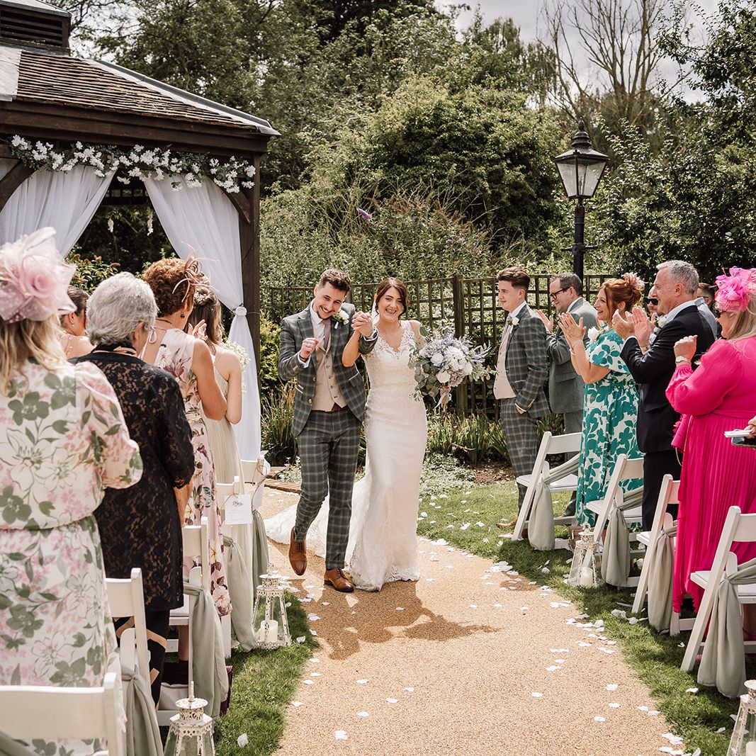A newlywed couple walks down the aisle outdoors beneath a decorated gazebo, smiling and celebrating as guests applaud on either side.