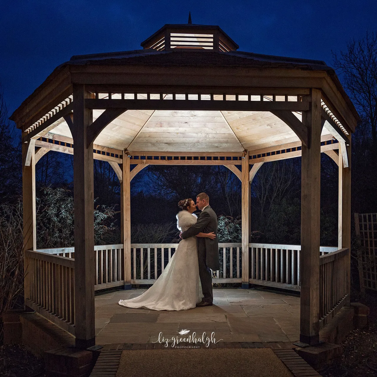 Bride and groom embrace in a beautifully lit wooden gazebo at night.