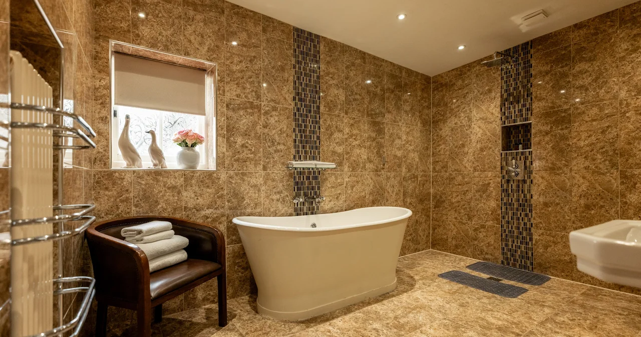 Luxurious bathroom with brown marble tiles, freestanding bathtub, window with decorative vases and flowers, a chair with folded towels, and modern fixtures.