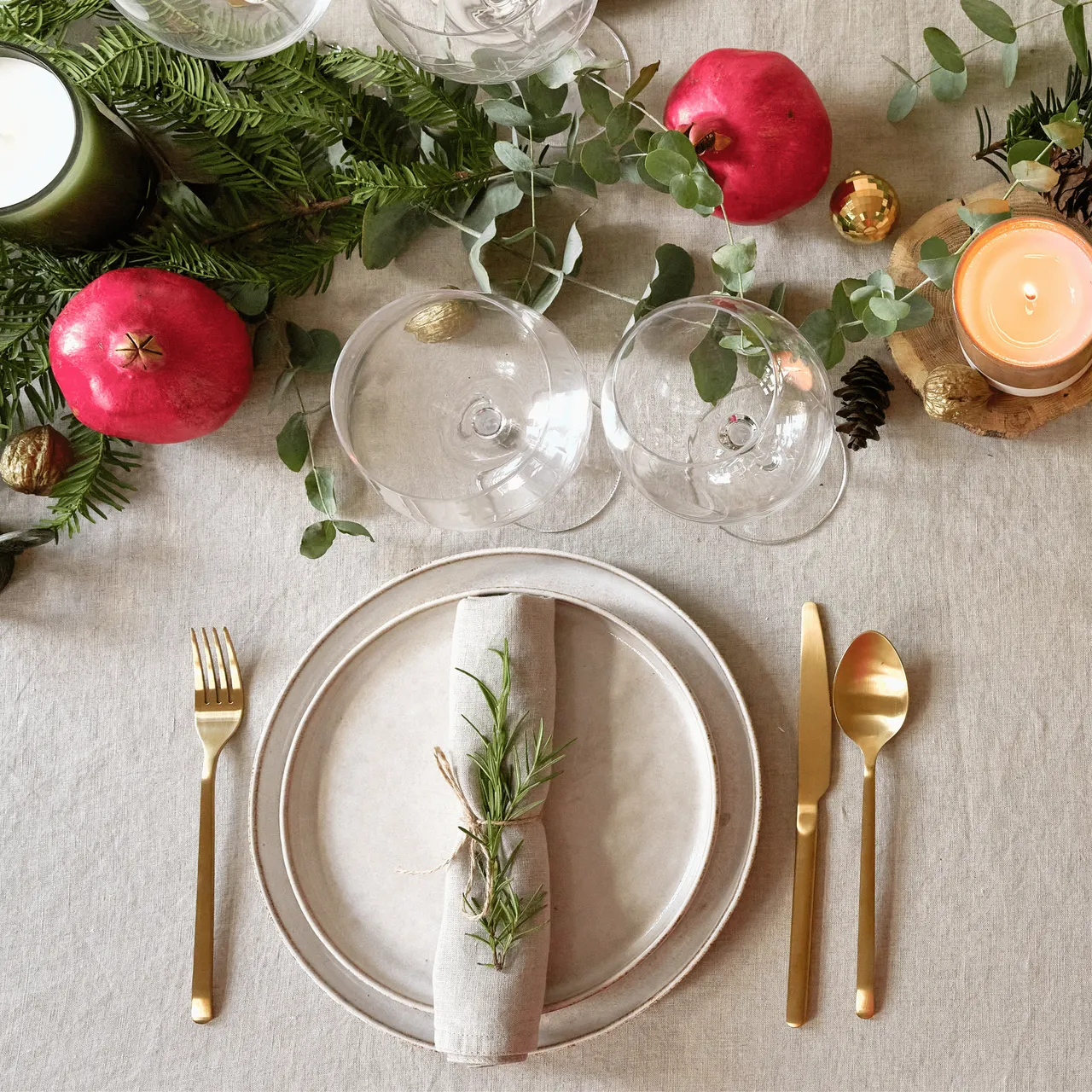 Elegant holiday table setting with gold cutlery, candles, greenery, pomegranates, and wine glasses.