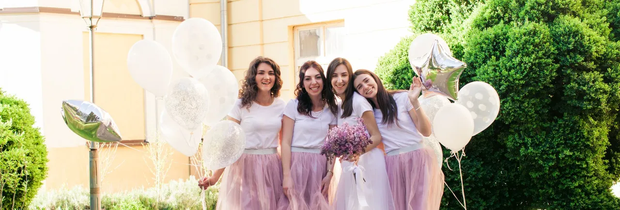 Bride and bridesmaids pose cheerfully outdoors, all wearing white tops and tulle skirts, holding balloons and flowers on a sunny cobbled path.