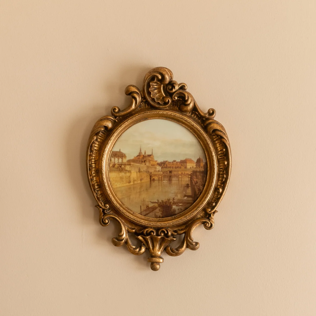 Ornate vintage gold-framed painting of a cityscape with river, hanging on a beige wall.