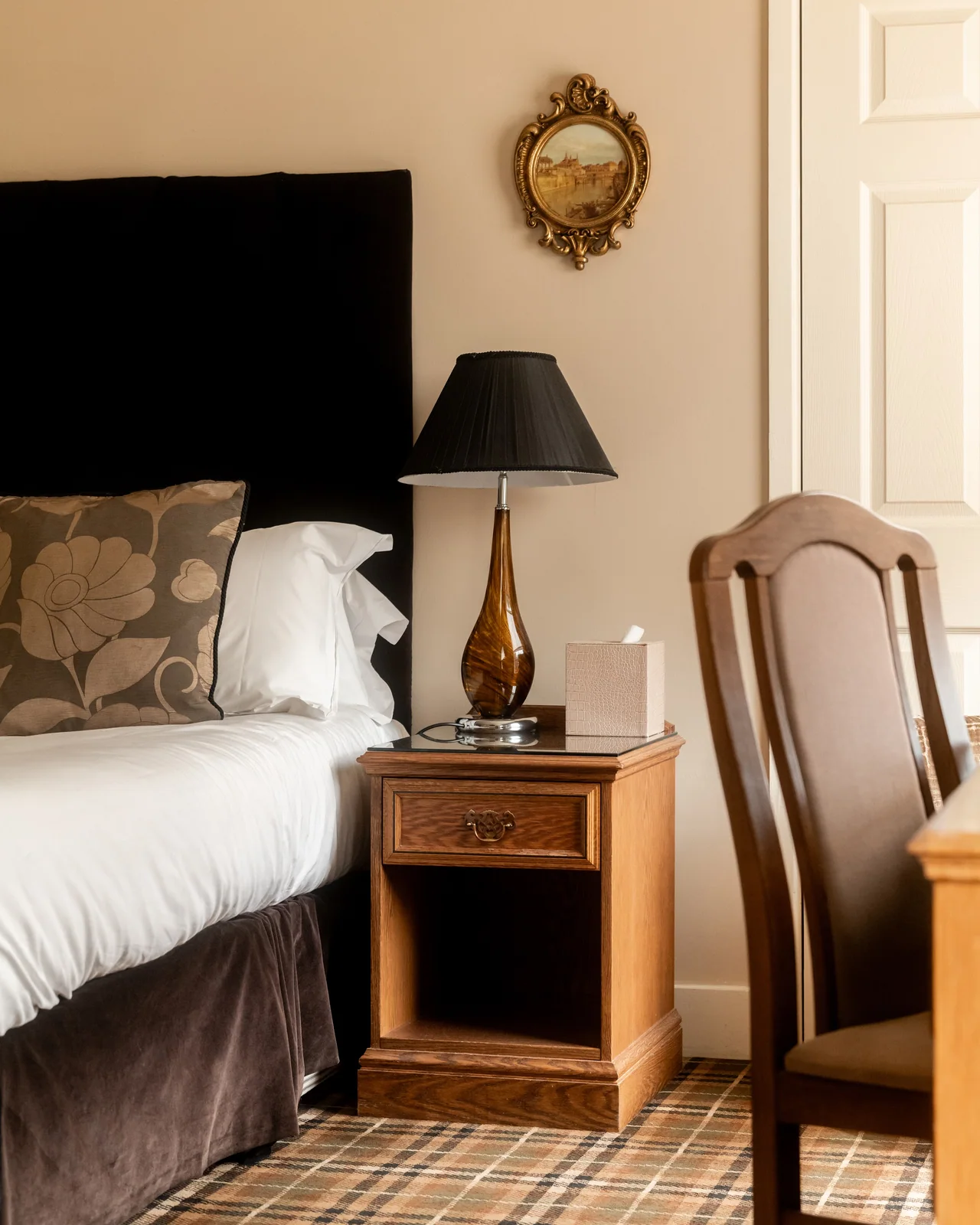 Cozy bedroom corner with a bed, floral pillow, wooden nightstand, brown table lamp, tissue box, and ornate framed painting on the wall.
