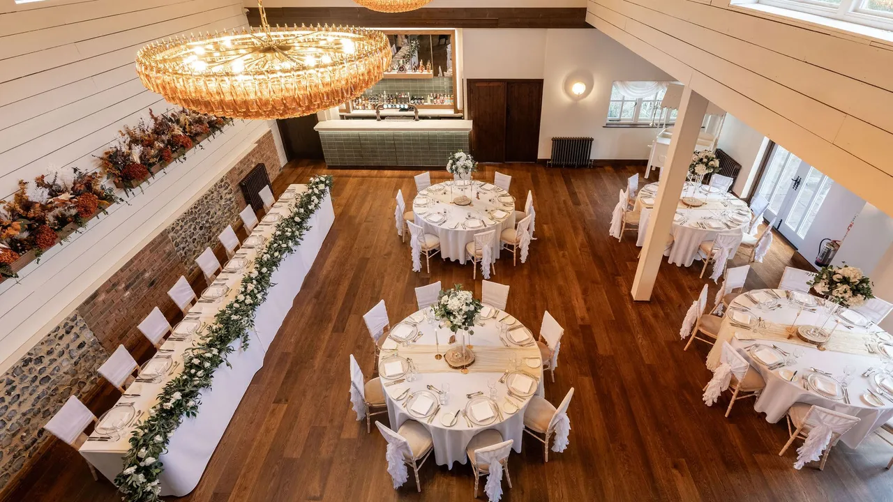 Elegant dining hall set up for an event with round tables, a long rectangular table, white chairs, and floral centerpieces.