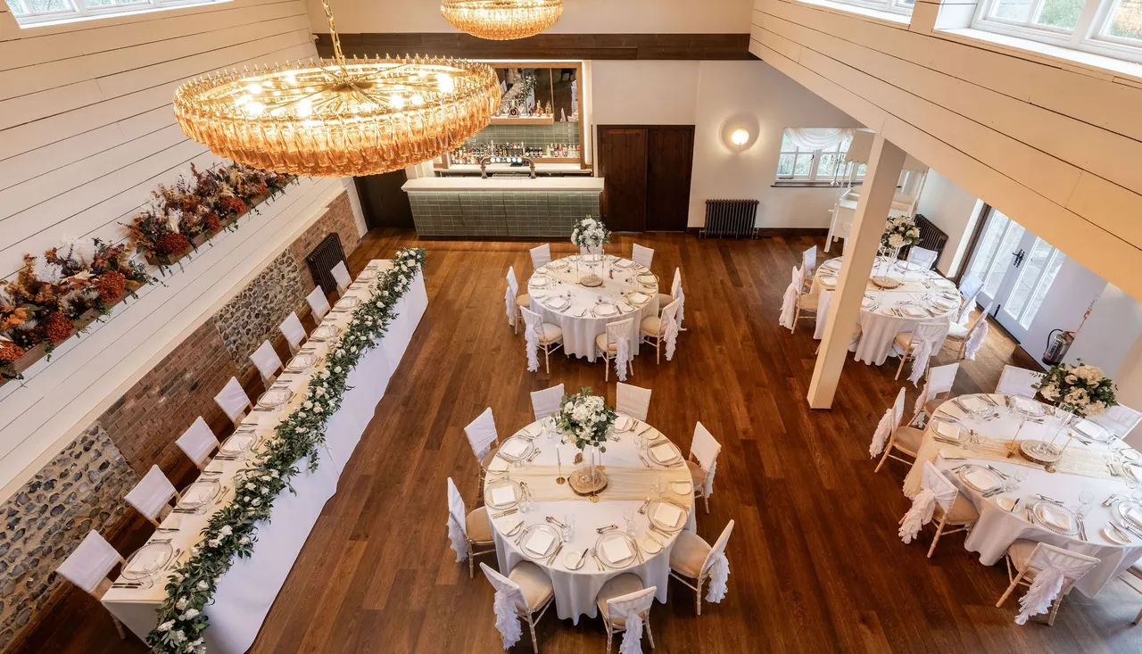 Elegant dining hall set up for an event with round tables, a long rectangular table, white chairs, and floral centerpieces.