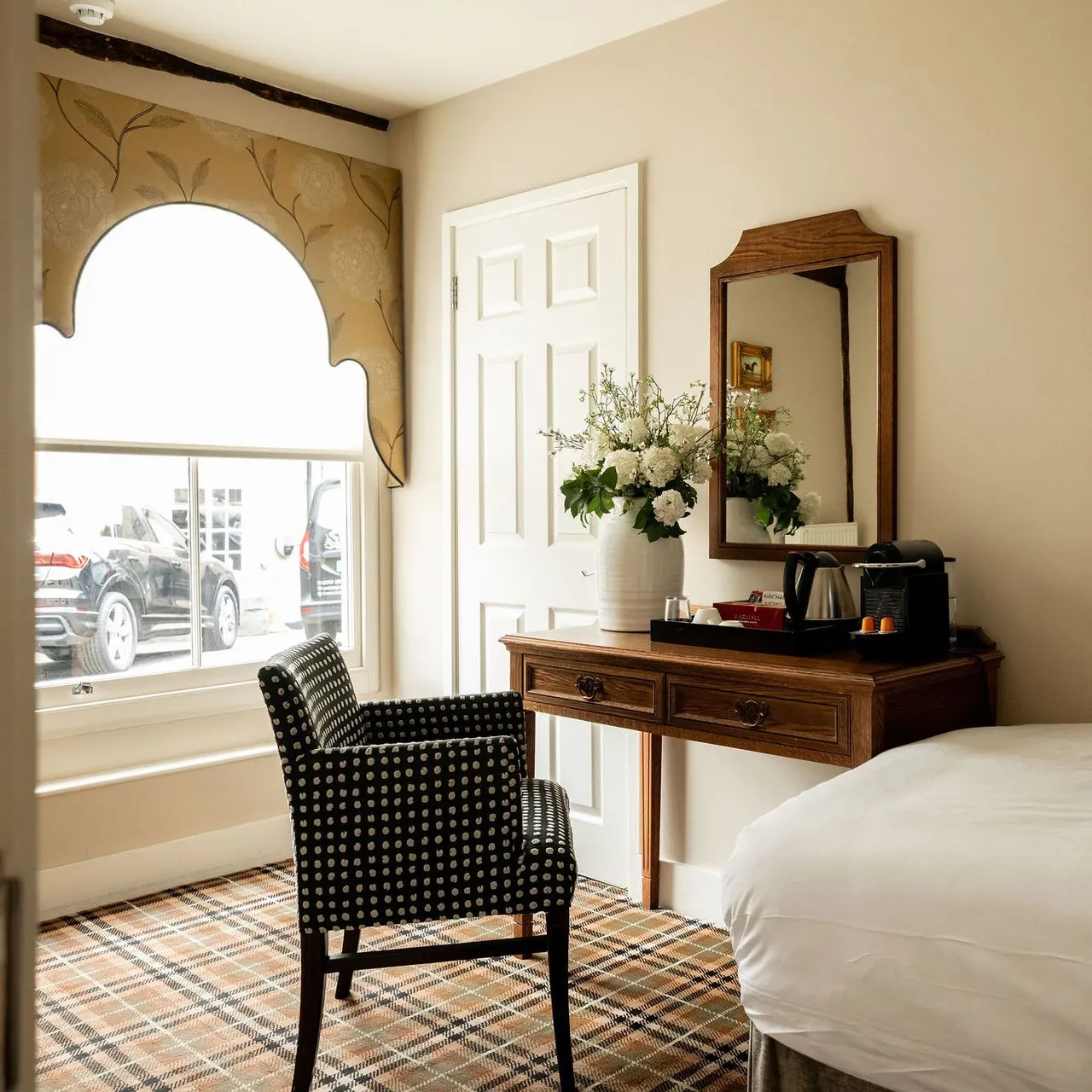 A cozy hotel room with a large window, patterned chair, wooden desk with mirror, vase of flowers, and coffee maker. The bed has white bedding and the floor is covered with a plaid carpet.