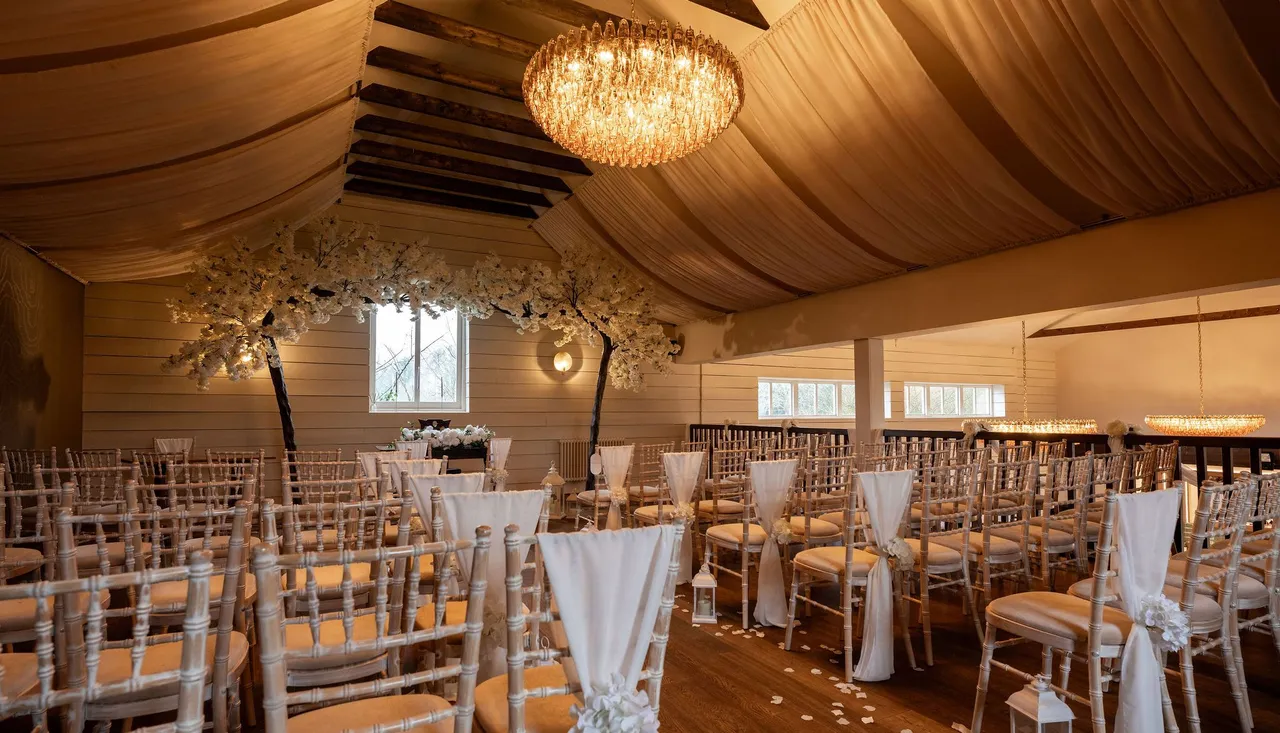 Elegant indoor wedding ceremony setup with rows of chairs, white drapes, flower arrangements, and chandeliers.