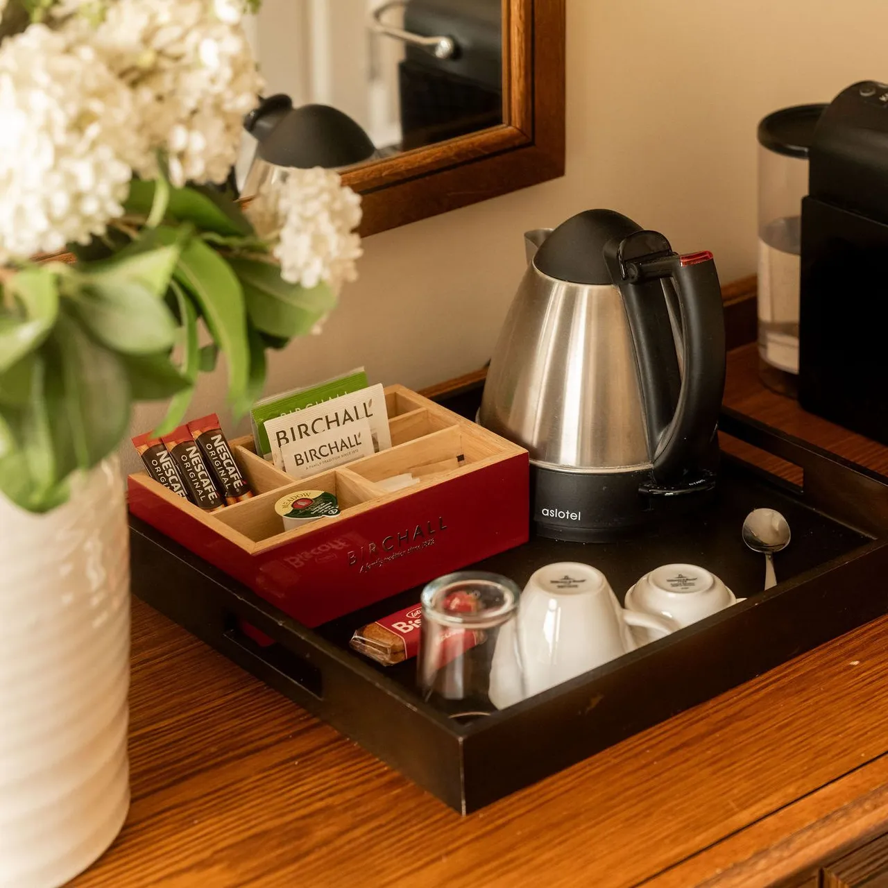 A hotel room refreshment tray on a wooden dresser with a kettle, Nespresso coffee machine with pods, tea, coffee sachets, mugs, glass, and a vase of white flowers.