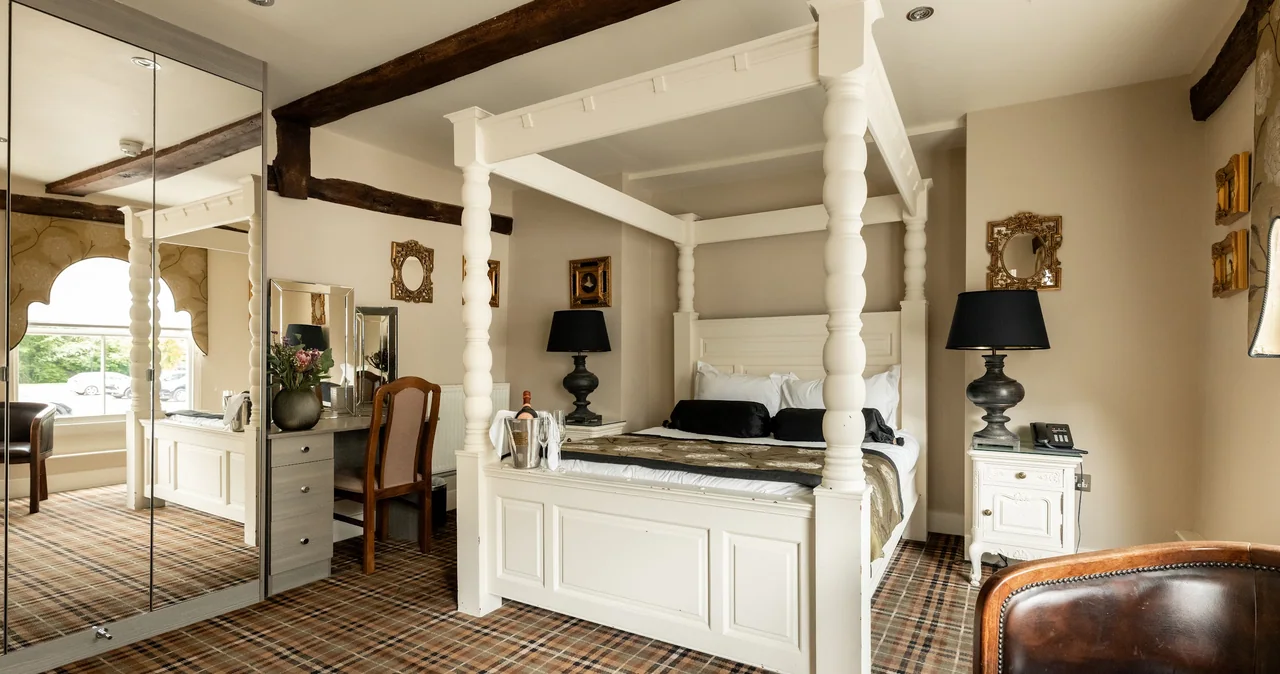 A well-decorated hotel room featuring a white four-poster bed, black lamps, a vanity with mirror and chair, plaid carpet, and classic framed mirrors on cream-colored walls.